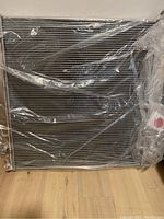 Front view of new aluminum radiator still wrapped in original plastic shipping protection, showing core and side tanks.