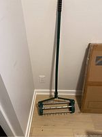 Full length image of green metal lawn air grader with long handle and wheeled roller with spikes at base against white wall and light wood floor.