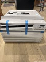 Front view of white LG window air conditioner with vent slats secured by blue tape, control panel with buttons and digital display.