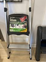 Photo of aluminum step ladder showing black plastic top platform with product label and rubber feet.