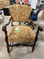 Front view of the antique chair showing carved wooden arms and floral upholstered seat and backrest.