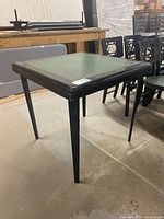 Square folding card table with green top and black legs shown standing on a concrete floor in a warehouse setting, angles visible of the legs and the frame top.