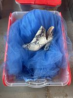 Plastic tote bin filled with assorted clothes and linens including visible blue tulle fabric and snake print boots.