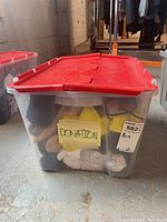 Plastic clear tote bin with red lid marked 'DONATION' with socks and shoes inside.