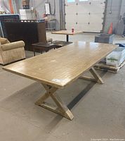 Photo showing the full top and one side of the rectangular wooden dining table with X-shaped base in a warehouse setting.