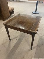 Square wooden end table with glass top, medium-dark brown finish, tapered legs ending in metal caps, visible wood grain, measuring about 26x26x21 inches.