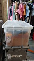 Clear plastic storage bin with locking lid containing assorted fabric pieces, placed on top of another storage bin with hanging clothes in the background.