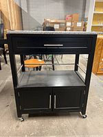 Front view of black MDF kitchen cart with speckled stone top, one drawer, open shelf, and cabinet doors below, on caster wheels.