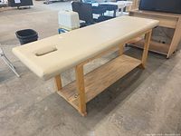 Full view of tan padded massage table bench with wooden frame and storage shelf underneath at warehouse.