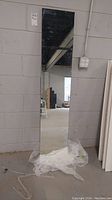 Full-length free-standing mirror leaning against a cinderblock wall with plastic wrapping at the base.