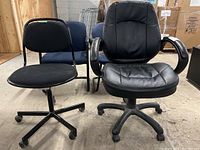 Two adjustable office chairs on casters placed in a warehouse environment, one black vinyl chair with armrests and one black fabric chair with metal tubular frame.