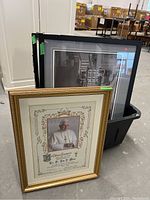 Three framed and matted prints in a bin, one gold frame with Pope print, two black framed photographic prints underneath, one with damaged frame.
