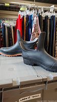 Pair of black rubber shoes displayed on top of a grey bin with clothes hanging in the background. Shoes have a mid-ankle height and orange-brown colored sole edges.