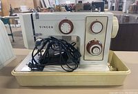 Side view of Singer model 5107 sewing machine in beige with brown dials, placed in its plastic carrying case with power cord coiled on it.