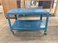 Front view of blue metal table cart with flat top and lower shelf, showing caster wheels and scratches on surface.