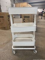 Front view of white metal utility cart showing three rectangular shelves and caster wheels.