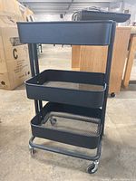 Front view of black metal utility cart with three tiers and caster wheels on concrete floor with wooden furniture and boxes in background.