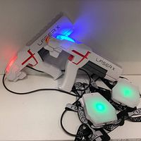 Two white Lazer X laser tag guns with red detail and two wearable sensor packs with green lights on straps and cables.