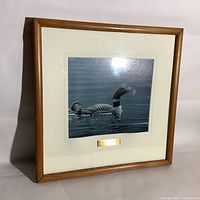 Framed artwork print of a loon with a chick on water, inside a light brown wooden frame with a plaque.