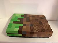 Front side view of Xbox One S console with Minecraft pixelated grass and dirt block design covering the top surface.