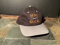 View of black and grey baseball hat, front showing embroidered text: Sens Suck with crossed golf clubs.