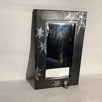 Front view of the black framed mirror with spiderweb decorations and metal hook at the bottom.