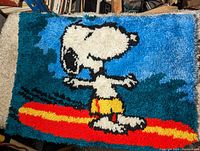 Full view of Snoopy latch hook rug showing Snoopy surfboarding in yellow swim shorts, with blue and green background.