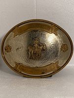 Front view of a vintage oval belt buckle with silver textured background, gold-tone borders and central cowboy on horseback figure.
