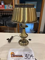 Front view of the brass electric table lamp showing the cord, rounded base, and tapered brass shade with finned top.