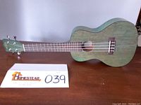 Full view of green Kala ukulele showing body and headstock with tuning pegs, resting on wood surface alongside auction lot card.