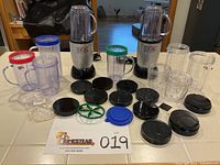 Two Magic Bullet MB1001 blender base units with multiple clear jars with different colored lids, flat and blade lids, and accessory parts spread out on a kitchen counter.
