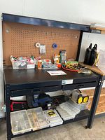 Full view of the black metal workbench with pegboard back, items on counter and lower shelf storage boxes and bags.