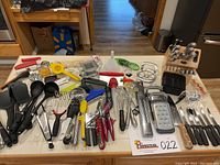 Full view of the kitchen utensils and tools laid out on table including KitchenAid spatulas, steel whisks, vegetable peelers, lemon squeezer, red handled tools, metal whisks, funnel, tongs, black ladles and turners, cutting knives, grater set and wooden knife block with knives.
