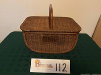 Front view of rectangular wicker picnic basket showing P&G emblem plaque on center of side with handle upright.