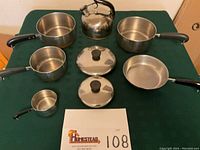 Full set of Revere Ware cookware displayed on green tablecloth, showing all items together: saucepans, skillet, tea kettle, measuring cup, and lids.