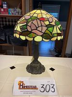 Image of rose stained glass lamp lit, showing pink rose designs with green, yellow, brown and red glass pieces on the shade, with ornate metal base and finial.