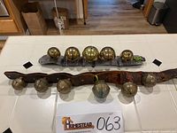 Two sets of vintage brass sleigh bells displayed on a tiled surface; one set mounted on a brown leather strap with six large bells, and another set of five smaller bells on a gray felt strap.