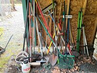 Photo showing a large assortment of yard tools leaning against a wooden wall inside a shed. Includes shovels, rakes, pitchforks, hoes, pruning loppers, a sprayer, and a green tripod stand. Ground is covered with dry leaves and soil.