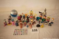 Wide view of the entire lot showing multiple children's toys including dolls and figurines arranged on a white backdrop, with a sheet of sticker decorations and a numerical lot card 108 visible.