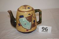 Photo showing full left side of majolica fan teapot with textured yellow base and greenish handle, multicolored fans, and floral decoration.