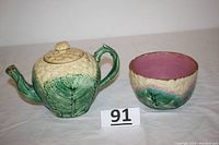 Wide shot of the Etruscan majolica teapot and bowl set with visible lid damage and chips on spout showing ceramic details and surface condition.