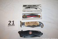 Photo showing three pocket knives including Fury Pocket Knife in box, NRA Elf Monkey (black handle), and Super Grip with wooden handle.