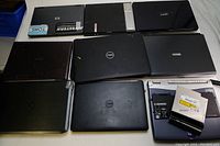 Top-down view of 10 assorted laptops closed, showing various brands and sizes, plus loose optical drives and components at the front.
