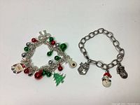 Two vintage Christmas charm bracelets laid on a white background. One bracelet has colorful small bells and multiple charms, the other has a simpler chain with three distinct Christmas charms.