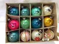 Top-down photo of 12 vintage mid century modern glass Christmas ornament balls arranged in a cardboard box with dividers. Various colors: blue, aqua, green, white, yellow, pink, and hand-painted floral designs. Some ornaments show paint peeling and discoloration.
