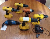 Five assorted battery-powered drills including four yellow and one orange, two chargers, and one blue corded multi-tool on a wooden table.