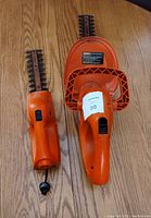 Two orange electric hedge trimmers showing main bodies and blades side by side on wooden surface.