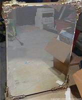 Full view of large rectangular mirror with gold decorative corners and frame, standing upright in a storage or warehouse area.