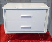 Front view of white side table with two drawers and metal handles, standing on metal legs on red fabric.