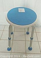 Side and partial angled view of shower stool showing round blue padded seat and silver metal adjustable legs with non-slip blue feet on tiled floor.